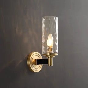 The Romea Copper Sconce by Morsale is a contemporary lighting fixture boasting a copper finish and a cylindrical glass shade that casts warm light on a gray textured wall. It features LED bulbs, accompanied by a circular base and vertical arm that supports the glass.