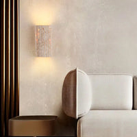 A modern living room featuring a beige sofa, a brown side table, and the Morsale Naples Travertine Sconce with LED bulbs emitting warm light, set against a neutral-toned wall with vertical gold accents.