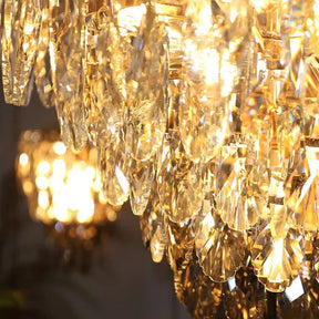 Close-up of the Lazzo Contemporary Chandelier by Morsale, showcasing its intricate glass design and the warm glow of LED bulbs. In the background, a smaller chandelier softly illuminates, enhancing the modern style and depth of this elegant setting. Text reads "LAZZO & DELUXE.