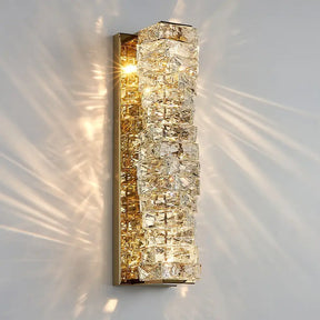 The Bacci Crystal Wall Sconces from Morsale.com feature a vertical, rectangular design with a brass frame adorned with multiple clear, angular crystal elements. The light shining through the crystals creates a dazzling starburst pattern on the surrounding wall, enhancing the elegant ambiance of this modern design.