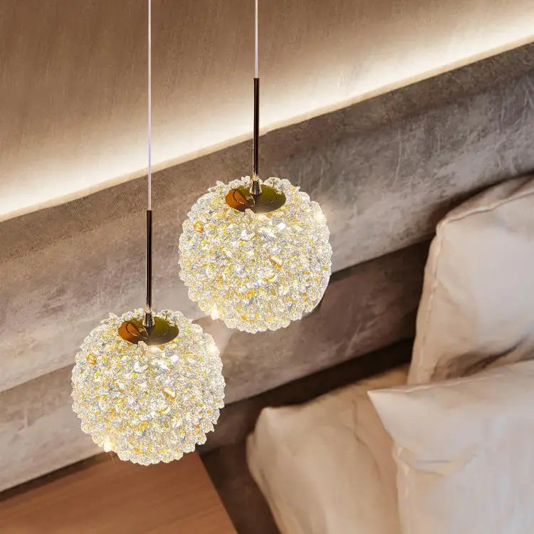 Two El Sol Crystal Pendant Lights from Morsale.com with intricate, sparkling designs hang above a modern bedside table. Crafted from premium materials, they cast an elegant illumination, offering a warm, ambient glow over a textured wall and a neatly made bed with white linens.