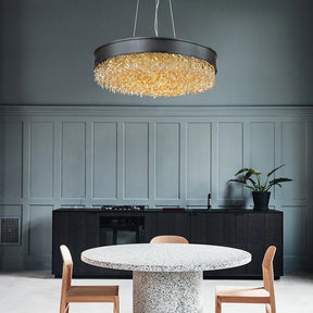 A modern dining room highlights the breathtaking Alleri Modern Chandelier by Morsale, casting its glow over a stone table. The area includes two wooden chairs, stainless steel accents, dark cabinetry, and a potted plant against light gray paneled walls.