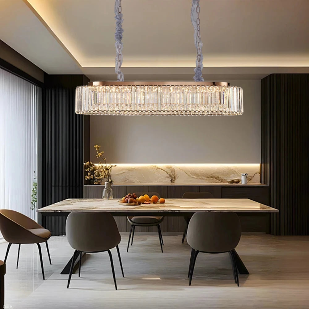 A modern dining room features a marble table surrounded by four chairs. Overhead, the Gio Crystal Dining Room Chandelier by Morsale elegantly illuminates the space. The back wall is adorned with dark cabinetry and illuminated shelves showcasing decorative items, while fresh fruit adds a natural touch.