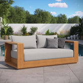 The Morsale Tahoe Acacia Wood Outdoor Patio Loveseat, featuring light grey cushions and a dark grey pillow, is set by the pool. Crafted with US-origin materials for quality assurance, it sits against greenery and trees beneath a blue sky with scattered clouds. Assembly required.