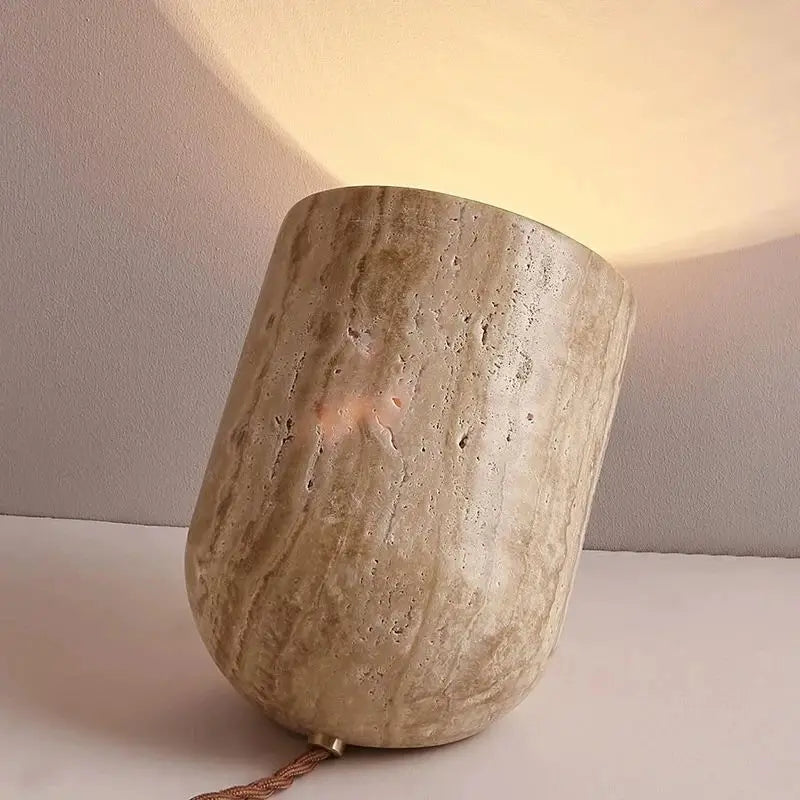 A small, cylindrical Modern Travertine Table Lamp from Morsale.com, with a concrete texture, tilts at an angle on a light-colored surface. Emitting a warm yellow light reminiscent of natural yellow cave stone, it casts a soft glow on the wall behind it. The power cord is visible trailing off to the side.