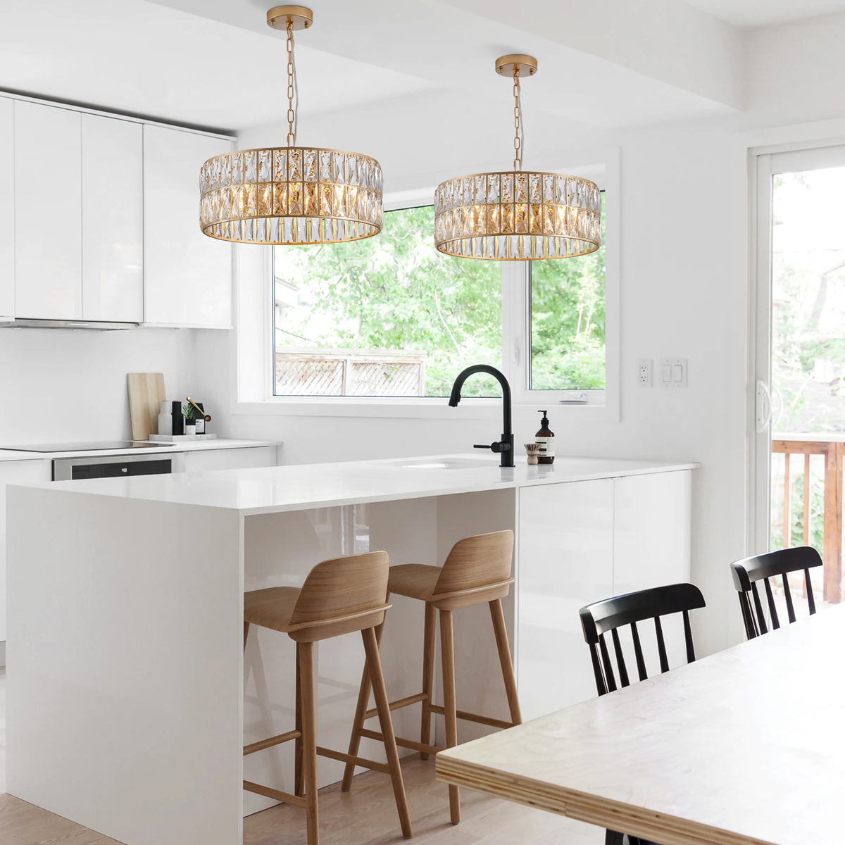 Modern white kitchen with a central island, two wooden adjustable bar stools, black faucet, dining table with black chairs, and large windows overlooking green trees. Illuminated by the Morsale Brion Crystal Kitchen Chandelier.