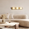 A modern living room features a light beige curved sofa, neutral pillows, a round wooden coffee table with dried grasses, and the Sonte Travertine Wall Sconce by Morsale.com—an architectural light with three glowing circular bulbs.