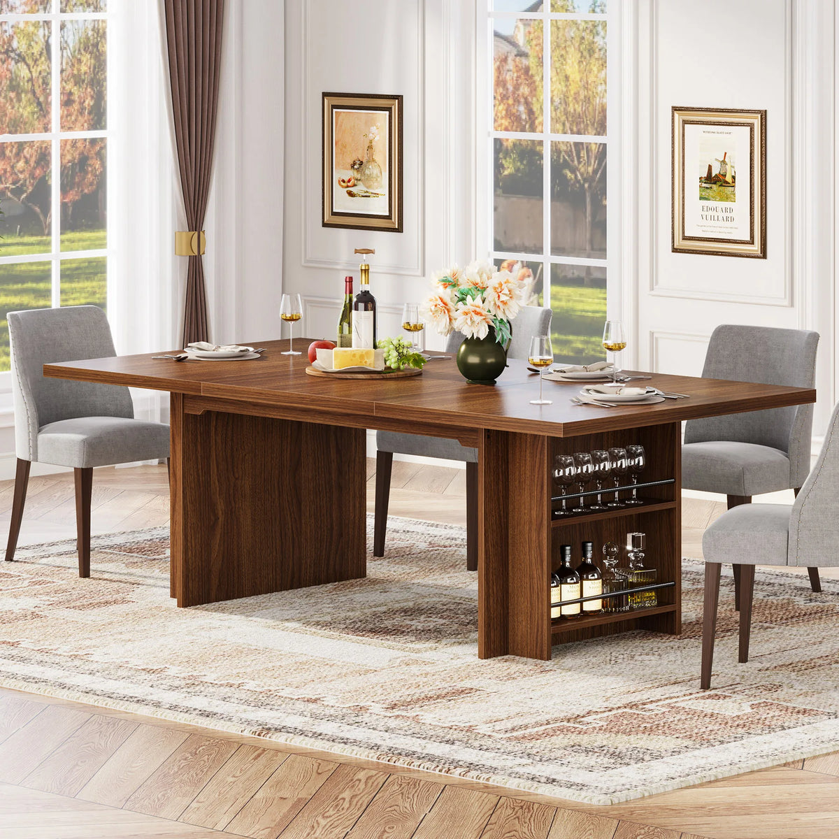 The Morsale Bristol 63" Dining Table with Storage is set with four gray chairs, wine glasses, a fruit platter, and a flower vase. A built-in shelf holds bottles and glasses. Large windows reveal a sunny day outside, and two framed pictures enhance the setting's charm.