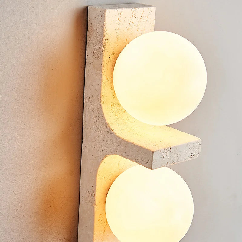 A modern Japan Style wall-mounted light fixture from Morsale.com, featuring two glowing circular LED bulbs, is vertically installed on a light-colored wall. Below the Natural Travertine Modern Wall Lights, to the left, a green leafy plant is partially visible.