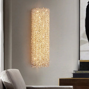 The Morsale Bergamo Crystal Sconce exudes a warm glow, lighting up a modern living room with its LED wall lamp. The space includes a beige armchair and a stack of books on a shelf, one of which has "Louis Vuitton" on the spine.