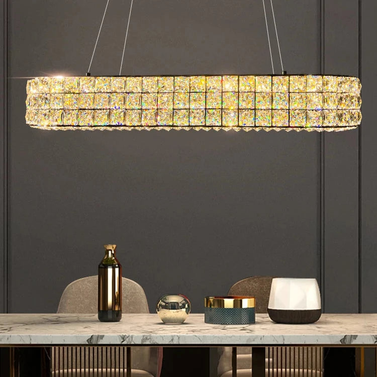 A luxurious dining area features the Bacci Crystal Dining Room Light Fixture by Morsale, elegantly illuminating a marble table with LED chandeliers. The setting contrasts against a dark wall with a gold bottle, glass sphere, and geometric containers.