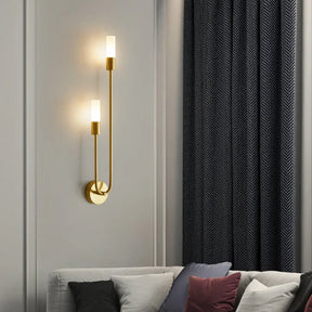 The Morsale Palermo 2-Light LED Wall Sconce, featuring two illuminated cylindrical glass shades, is stylishly installed on a gold fixture against a white paneled wall. Below, a sofa embellished with black, white, and red cushions enhances the modern aesthetic. A dark curtain with a subtle pattern can be partially seen on the right.