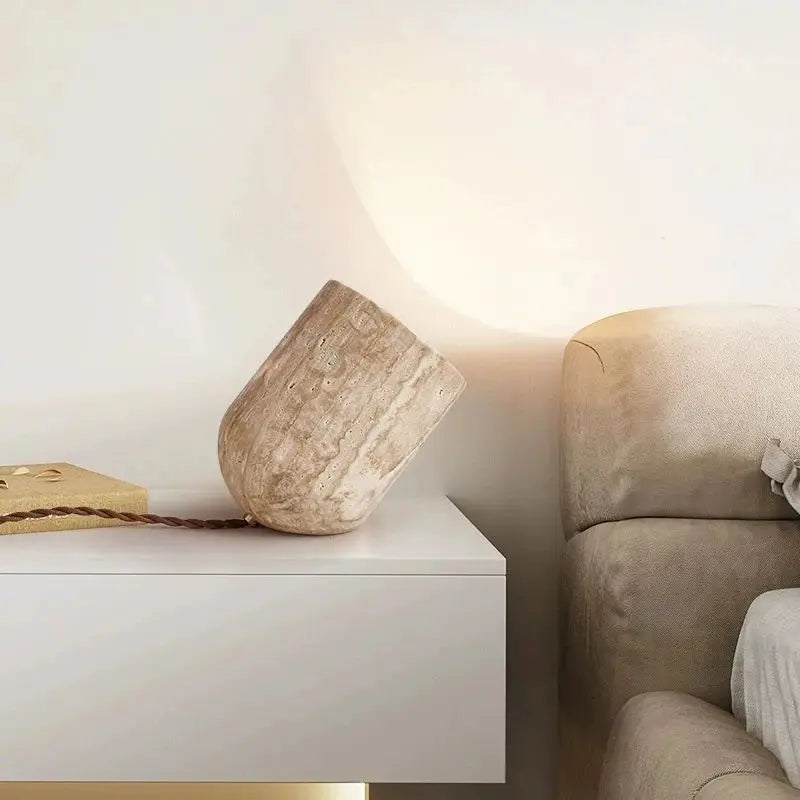 A small, cylindrical Modern Travertine Table Lamp from Morsale.com, with a concrete texture, tilts at an angle on a light-colored surface. Emitting a warm yellow light reminiscent of natural yellow cave stone, it casts a soft glow on the wall behind it. The power cord is visible trailing off to the side.