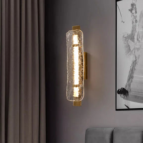 A Copper & Glass Modern LED Wall Sconce by Morsale, featuring a cylindrical design with textured glass and warm LED lighting, is installed on a gray wall. Nearby are elements like hints of an abstract black and white painting and dark curtains, enhancing the ambiance.