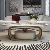 A modern living room features a 55" Morsale round marble cocktail table with storage and a gold base, suggesting US craftsmanship. A white potted plant sits atop, complemented by a gray sofa with a red cushion, abstract wall art, and an elegant dining area in the background.