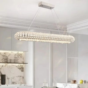 A modern kitchen showcases a Morsale Bacci Crystal Modern Chandelier with LED bulbs, casting a gentle glow from the ceiling. Marble backsplash and countertops, light gray cabinets, and glass shelves enhance the elegant and contemporary style of the space.