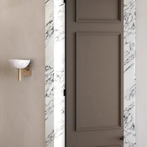 A minimalist interior showcases a closed taupe door framed by exquisite white Spanish marble with black veins. Adjacent to the door, mounted on the light gray wall, is the Morsale Spanish Marble & Copper Antique Sconce, featuring a contemporary design with a white half-sphere shade and gold base.