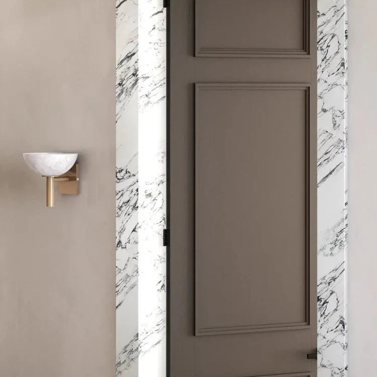 A minimalist interior showcases a closed taupe door framed by exquisite white Spanish marble with black veins. Adjacent to the door, mounted on the light gray wall, is the Morsale Spanish Marble & Copper Antique Sconce, featuring a contemporary design with a white half-sphere shade and gold base.