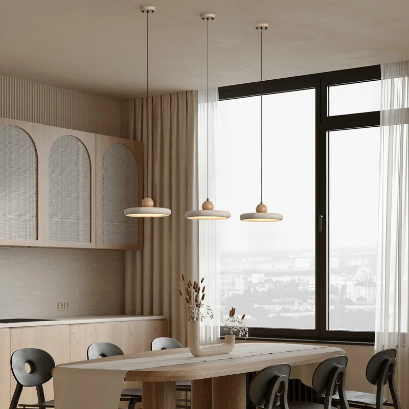 A minimalist dining area featuring a small table with dark legs and a light wooden top, surrounded by four chairs. The beige walls are adorned with a black-and-white abstract framed picture. Overhead, modern Natural Travertine Kitchen Pendant Light Fixtures from Morsale.com fitted with LED bulbs illuminate the space, which is subtly decorated with small items.