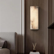 The Morsale Natural Marble LED Wall Sconce, featuring a modern cylindrical design, emits a warm glow on a wooden-paneled wall beside a gray sofa. Below it sits a small table adorned with books and a reed diffuser, enhancing the contemporary ambiance.