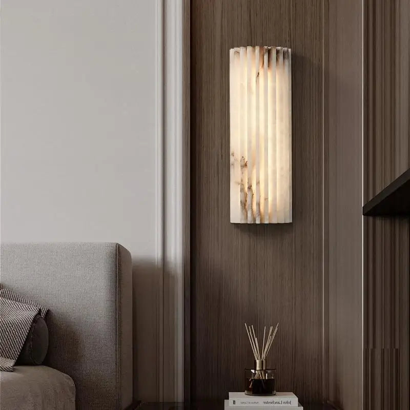 The Morsale Natural Marble LED Wall Sconce, featuring a modern cylindrical design, emits a warm glow on a wooden-paneled wall beside a gray sofa. Below it sits a small table adorned with books and a reed diffuser, enhancing the contemporary ambiance.