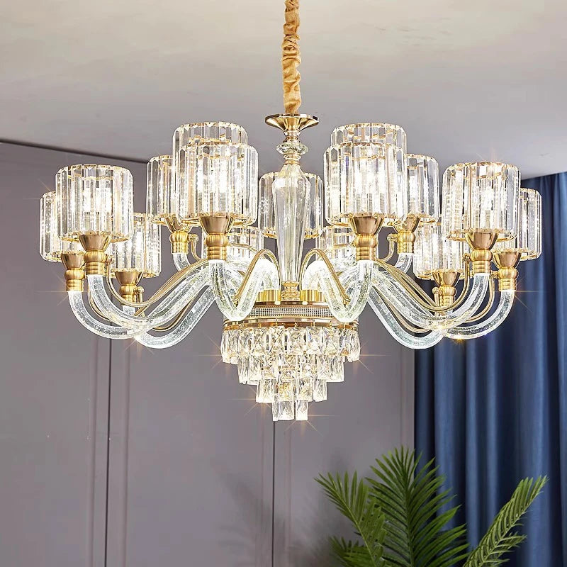 The Gio Crystal Contemporary Chandelier by Morsale is a luxurious piece featuring a gold frame with multiple glass arms, each hosting a cylindrical LED bulb. Its base is adorned with cascading crystal prisms, set against a light gray wall and complemented by a blue curtain and hints of green plant leaves.