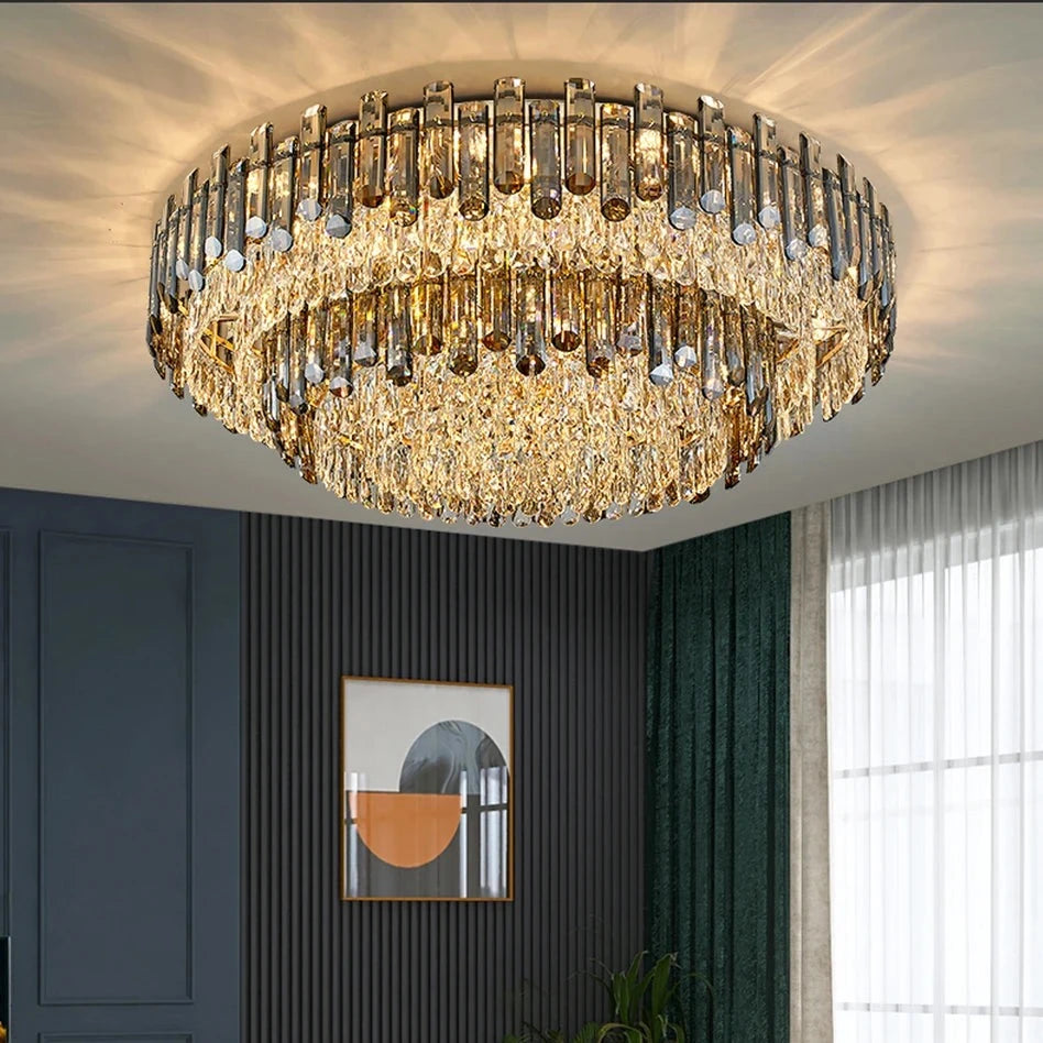 A luxurious living room features a classic luxury lighting fixture with the Venice 2-Tier Crystal Chandelier by Morsale.com adorned with cascading crystals. The room boasts modern furniture, including a beige sectional sofa with colorful cushions, a round glass coffee table, and a dark blue accent wall adorned with geometric art. Tall green curtains frame a large window.