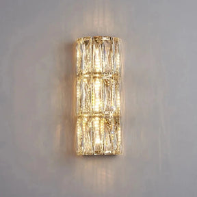 The Bacci Crystal Wall Lights by Morsale.com feature a modern design with a cylindrical shape and a lattice of metallic and handmade crystals. When switched on, they emit a warm and inviting glow against a plain, light-colored wall.