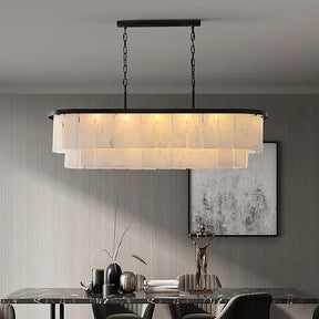 In a modern dining room, Morsale's Moonshade Marble Chandelier, an elegant oval fixture with translucent panels and a marble shade, hangs above a glass table set with decorations. The wall features a monochromatic abstract painting.