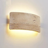 The Natural Travertine LED Wall Light Fixture by Morsale.com is an interior wall sconce featuring a rectangular, curved stone-like cover that emits a warm, soft glow. This energy-efficient LED lighting creates a halo effect on the white wall, emphasizing the texture and natural imperfections of the stone surface.