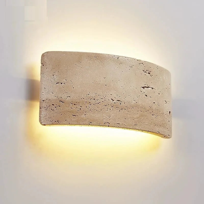 The Natural Travertine LED Wall Light Fixture by Morsale.com is an interior wall sconce featuring a rectangular, curved stone-like cover that emits a warm, soft glow. This energy-efficient LED lighting creates a halo effect on the white wall, emphasizing the texture and natural imperfections of the stone surface.