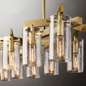 The Leroi Brass Dining Room Chandelier by Morsale showcases a modern design with a gold-colored metal frame and multiple cylindrical glass shades. The textured, rippled appearance of the glass encases visible filament light bulbs that emit a warm glow, all set against a gradient gray background.