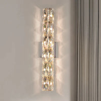 The Bacci Crystal Wall Sconce by Morsale.com emits a warm glow, creating intricate reflections on light-colored walls near tall curtains, enhancing your indoor lighting with its vertical, handmade crystal design.