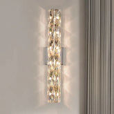 The Morsale.com Bacci Indoor Crystal Wall Sconce features a stylish, handmade rectangular crystal design that emits warm light. Mounted on a light-colored wall next to beige curtains, the elegant sconce enhances the ambiance. Below it, green palm leaves are partially visible, casting intriguing shadows on the wall.