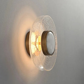 The Art Deco Cracked Glass Wall Sconce by Morsale is a contemporary light fixture that features a round, crackled glass shade with a textured surface. It includes a central, circular metal piece finished in brass and casts a warm glow on the wall to add an Art Deco flair.