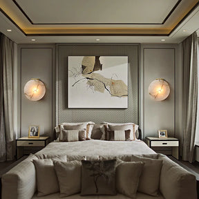 A stylish bedroom showcases a large bed with neutral bedding and abstract wall art. Two matching bedside tables featuring framed photos are flanked by Morsale's Moonshade Marble Wall Lamps in a modern style. Soft, flowing curtains frame the tall windows, infusing contemporary elegance.