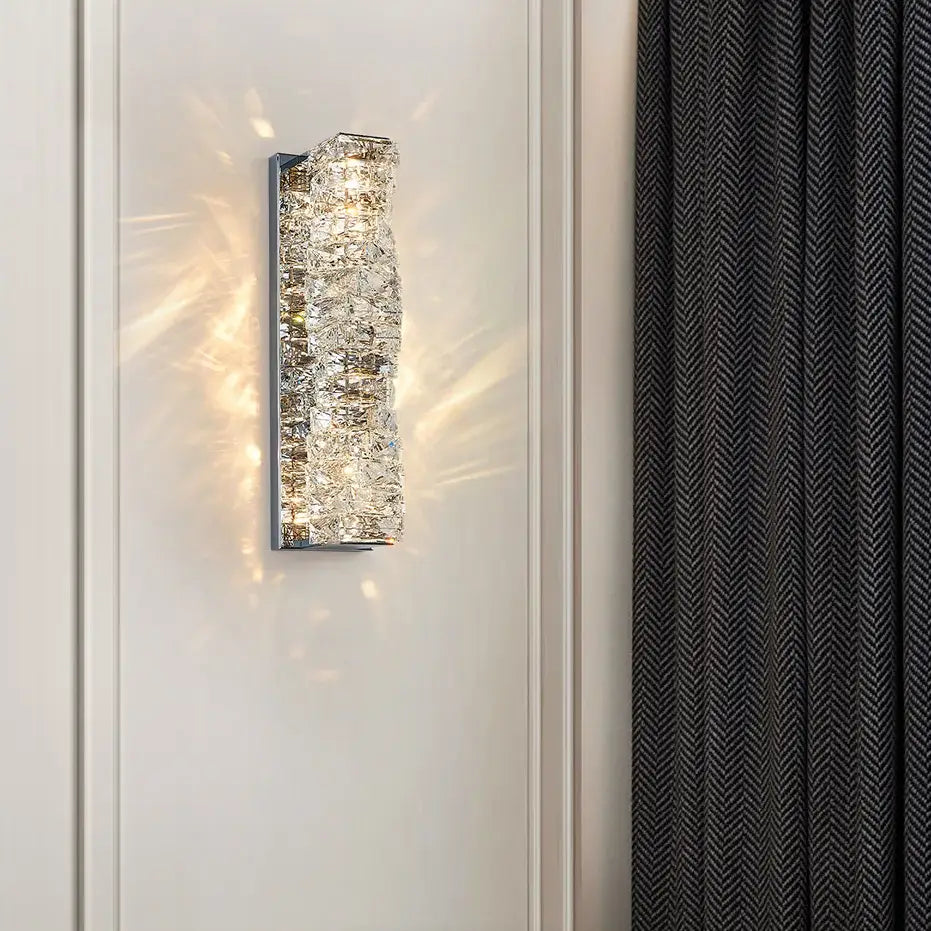 The Bacci Crystal Wall Sconce from Morsale.com, featuring a shimmering, textured metallic finish and a brass frame, emits a warm light. Mounted on a white wall next to a dark, textured curtain, it casts intricate light patterns on the surrounding area.