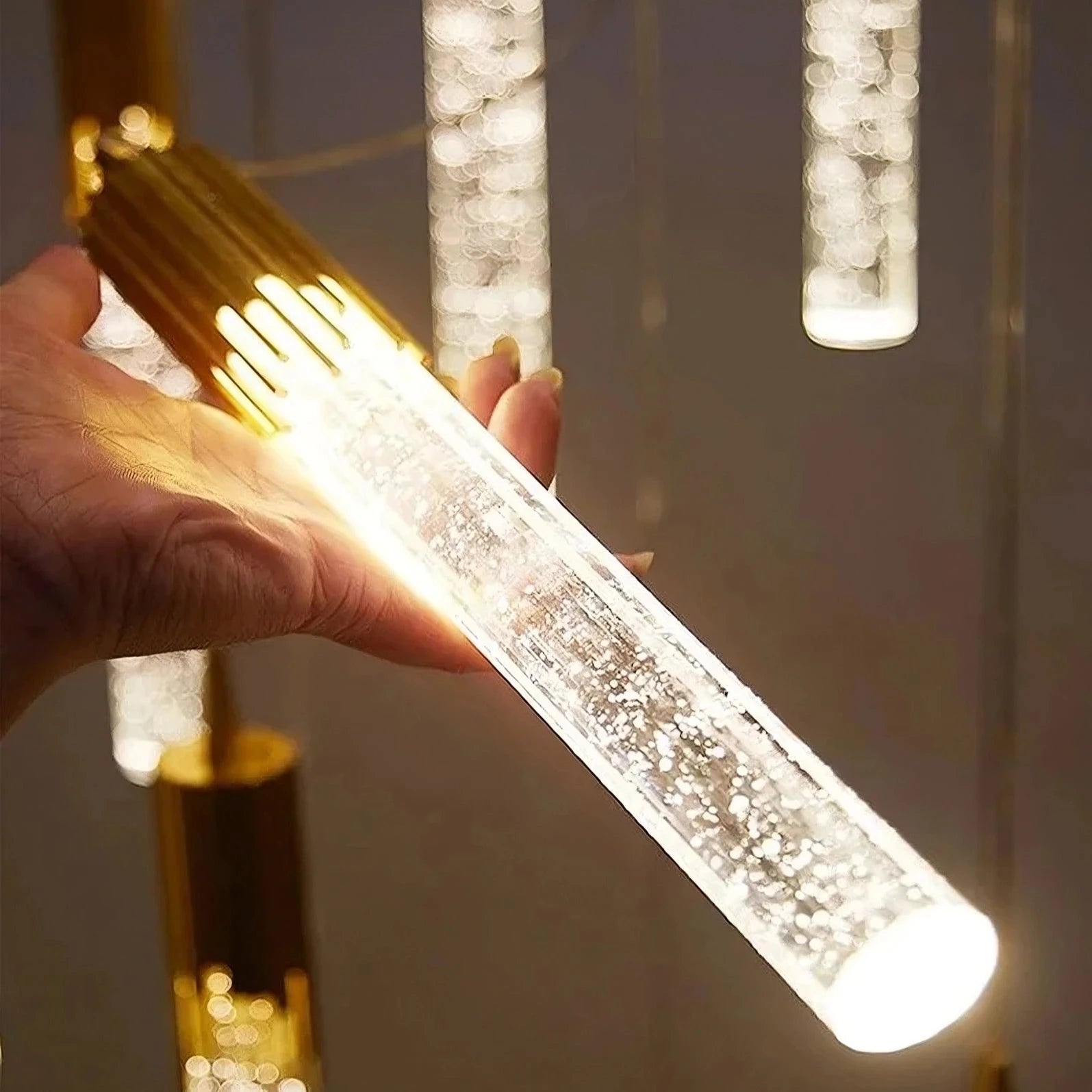 A person holds a glowing, cylindrical pendant light fixture made of K9 bubble crystal, with a polished gold finish that adds elegance. Several similar dimmable Modern Minimalist LED Pendant Lights hang in the background. The top text reads "Warm and Elegant Style," with "Product Details" in a black box. Brand Name: Morsale.com
