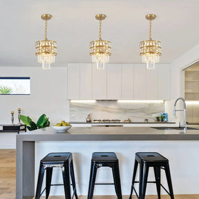 A modern kitchen with a sleek gray island featuring three black stools. Elegant Gio Crystal Kitchen Pendant Lights by Morsale, in luxurious gold and crystal, hang overhead, adding a touch of flair. White cabinetry and a marble backsplash complete the contemporary design.