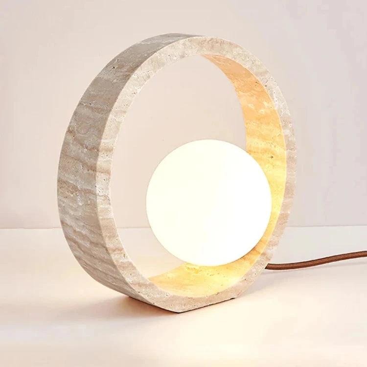 The Modern Travertine LED Table Lamp by Morsale.com features a round Natural Yellow Cave Stone frame with a textured, natural finish and a glowing spherical light bulb in the center. The lamp emits a warm, ambient light, creating a cozy atmosphere, and has a cord extending from the back.