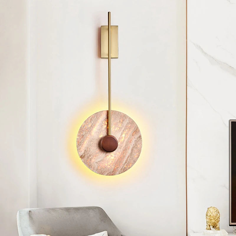 The Moonshade Natural Marble Sconce by Morsale casts a warm light against the textured wall with its modern circle design. It features a sleek vertical gold rod and round element, bringing Japanese-style minimalism to the space. The LED wall lamp enhances the ambiance, while a gray chair rests partially visible in the foreground.