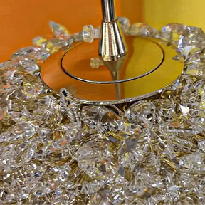 A close-up shot of a shiny metallic object, confirmed as the base of an El Sol Crystal Pendant Light by Morsale.com, adorned with numerous clear, faceted crystals that resemble diamonds. The crystals are densely packed, reflecting light and creating an elegant illumination. A polished silver rod extends from the center.



