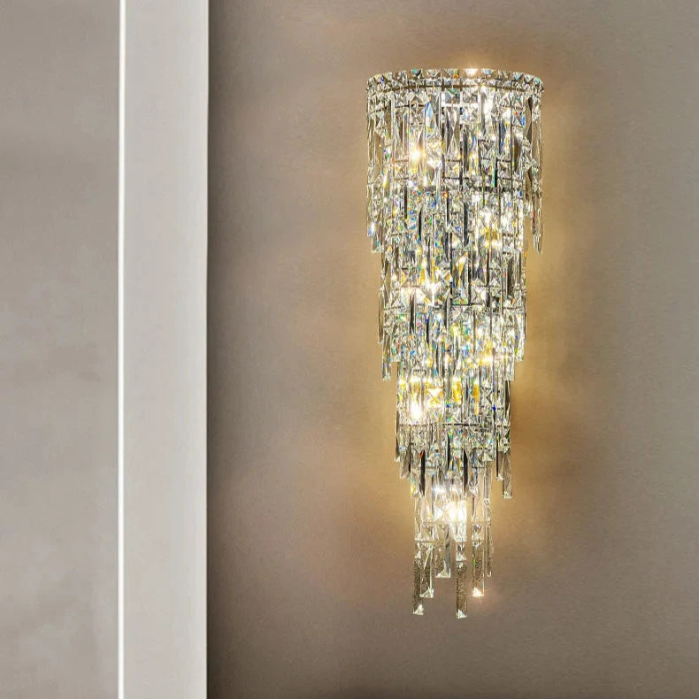 The Bacci Crystal Contemporary Wall Sconce by Morsale emits a warm, radiant glow with its wall-mounted design that features multiple cascading layers of handmade reflective crystal pieces, creating an elegant and luxurious atmosphere against a plain, light-colored wall.