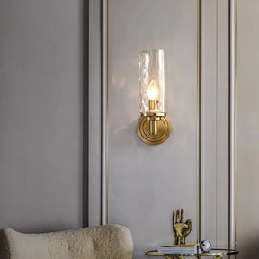 The Romea Copper Sconce by Morsale softly illuminates the gray paneled wall with its copper base and elongated glass shade. Below, a small table displays a sculpted gold hand and a round white decorative item. A beige chair is partially visible, subtly highlighting the presence of LED bulbs.