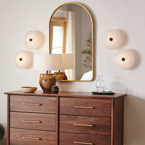 A wooden dresser with gold handles features a lamp, decorative bowl, and glassware. Above it hangs a large oval mirror with a gold frame, flanked by four Morsale Witney Copper & Marble Wall Sconces. The modern style adds elegance as the mirror reflects a window with a white curtain.