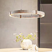A modern dining room features a round wooden table, a ceramic vase with pink and white flowers, and a Morsale Natural Travertine Modern Light Fixture with LED bulbs overhead. A textured wall and vertical glass panel in the background complete the contemporary look.