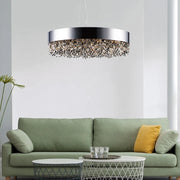 A modern living room showcases a green sofa adorned with yellow and gray pillows. In front, a round metallic coffee table is topped with glassware. Overhead, the Alleri Modern Chandelier by Morsale with crystal details casts an elegant glow throughout the space. A tall plant stands to one side, bringing in a natural element.