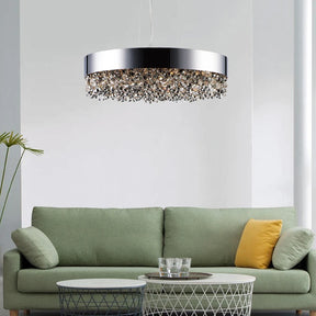 A modern living room showcases a green sofa adorned with yellow and gray pillows. In front, a round metallic coffee table is topped with glassware. Overhead, the Alleri Modern Chandelier by Morsale with crystal details casts an elegant glow throughout the space. A tall plant stands to one side, bringing in a natural element.