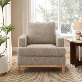 A Morsale Oversized Mid Century Modern beige armchair with wooden legs sits on a striped area rug, accompanied by a wooden side table holding a lamp and decor items. A large window showcases greenery outside, while a tall plant nearby adds to the room's cozy ambiance.
