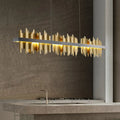 A modern kitchen with a concrete island is illuminated by the Rivaro Linear Chandelier from Morsale.com, featuring gold, jagged-edged panels that cast a warm glow and serve as a stunning artistic accent over the minimalist space.